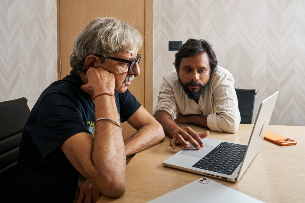 Ritesh Sharma collaborates with a fellow crew member at a desk, looking at a MacBook Pro. iPhone 17 Pro Max sits on the desk next to them.
