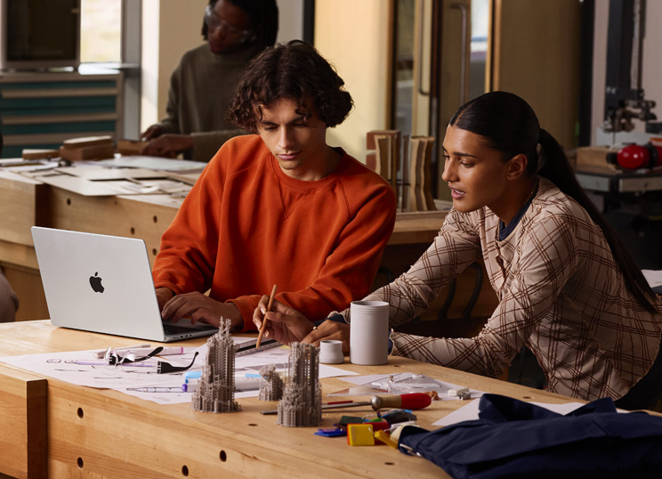 Deux personnes travaillant ensemble sur un projet à une table : l’une tape sur un MacBook, l’autre utilise un crayon et une règle sur du papier, des objets d’art les entourent.